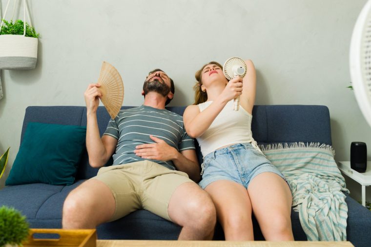Exhausted couple lounging on a sofa at home, battling summer heat wave with handheld and electric fans while sweating in discomfort