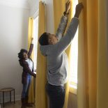 An African American stand near the bedroom windows, closing the blinds, while wearing casual clothing.