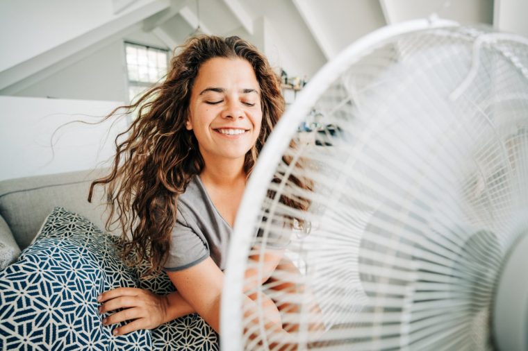 Happy contented woman using electric fan at home enjoying fresh air on hot summer day.