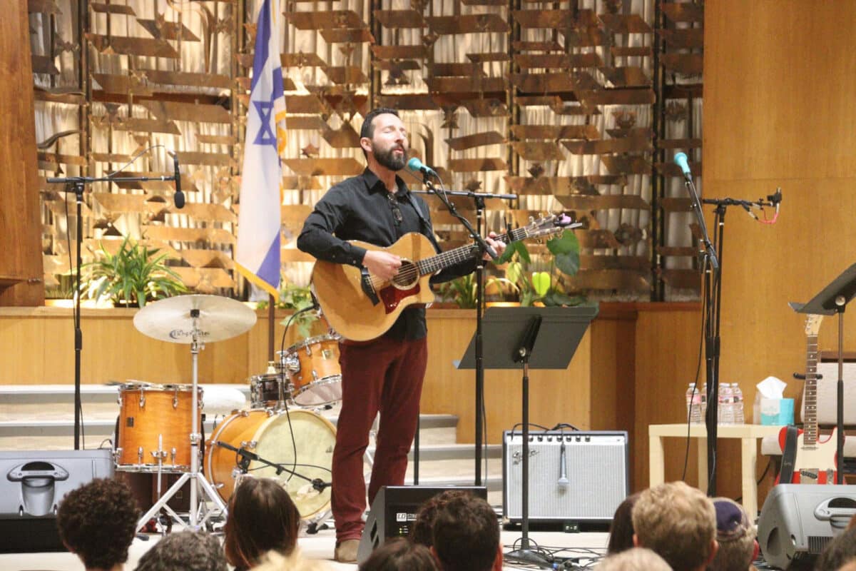 Alameda Post - Saul Kaye stands at the front of a room of worship with a guitar. 