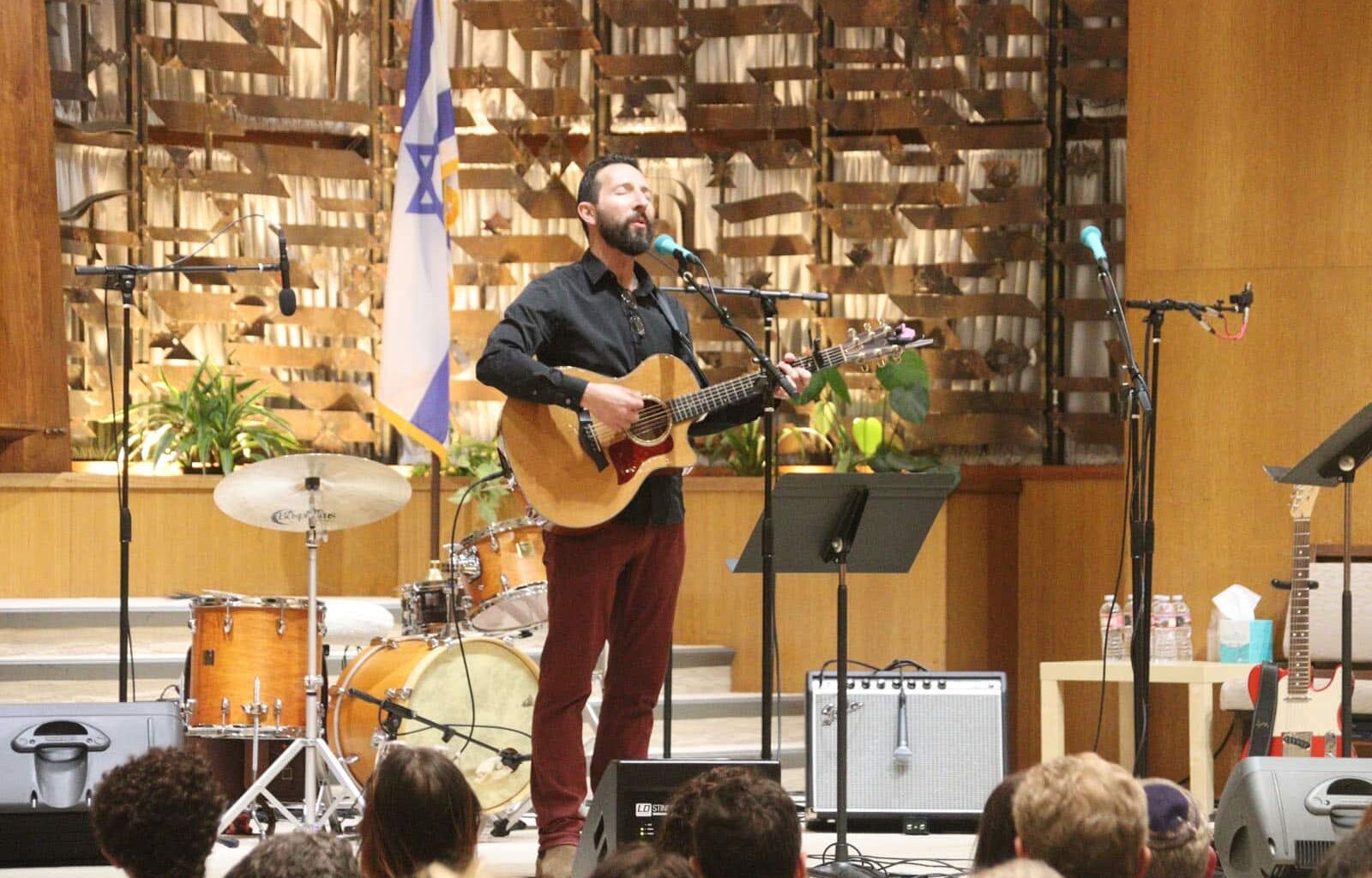 Alameda Post - Saul Kaye stands at the front of a room of worship with a guitar.