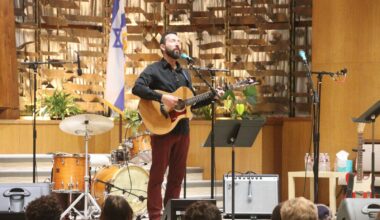 Alameda Post - Saul Kaye stands at the front of a room of worship with a guitar.
