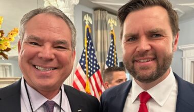 Two men in suits smiling for a selfie with American flags and a formal room backdrop behind them accessibly in the background.