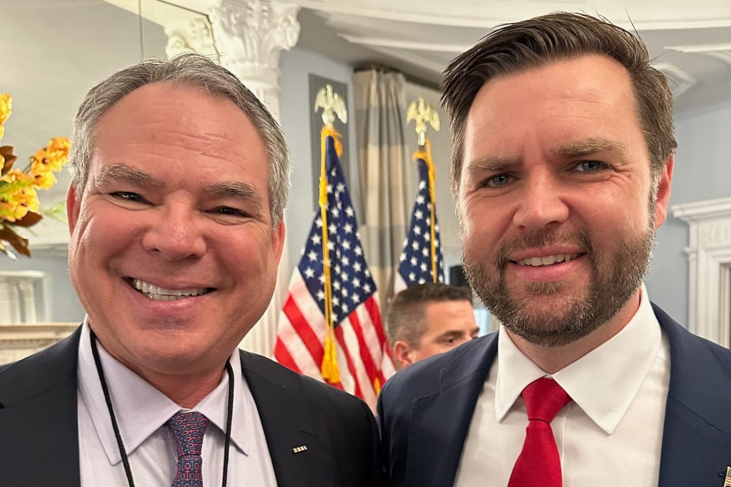 Two men in suits smiling for a selfie with American flags and a formal room backdrop behind them accessibly in the background.