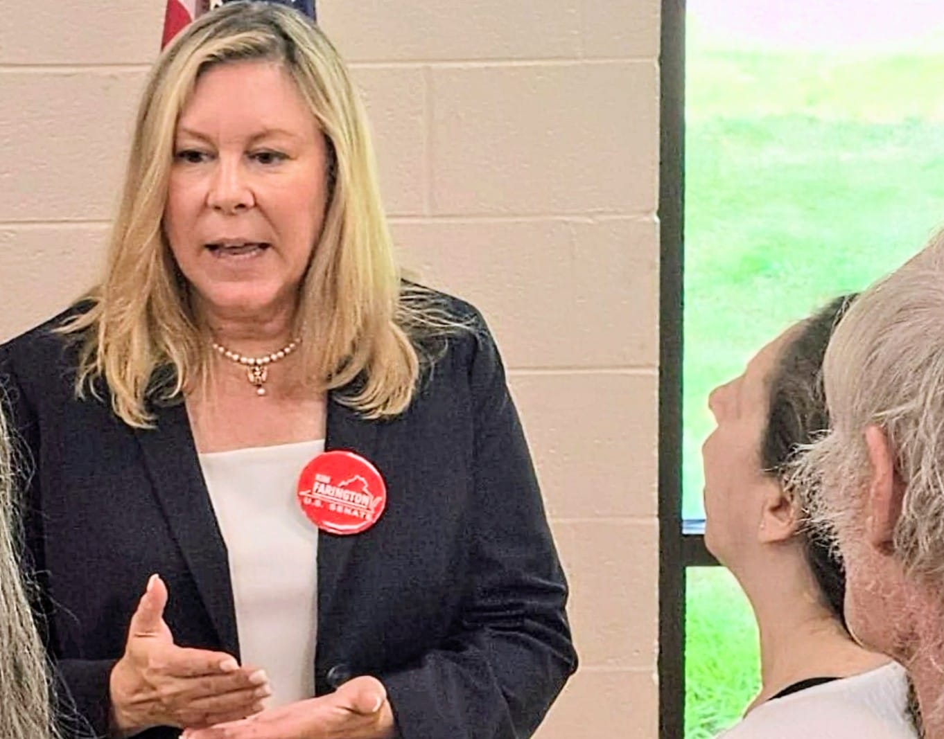 Woman in a black blazer speaks to a group, wearing a red campaign button on her lapel.