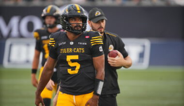 A football player wearing a black and yellow Tiger-Cats jersey with the number 5 walks on the field, accompanied by a coach holding a football.