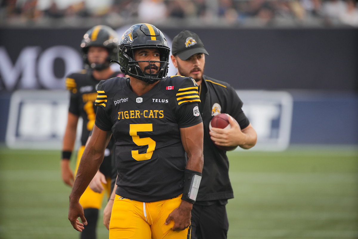 A football player wearing a black and yellow Tiger-Cats jersey with the number 5 walks on the field, accompanied by a coach holding a football.