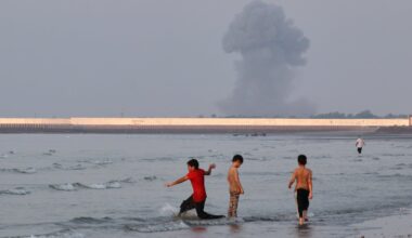 Three boys play in the shallow waters of the Strait of Hormuz, as a plume of smoke rises from an explosion in the background, off Bandar Abbas, Iran.