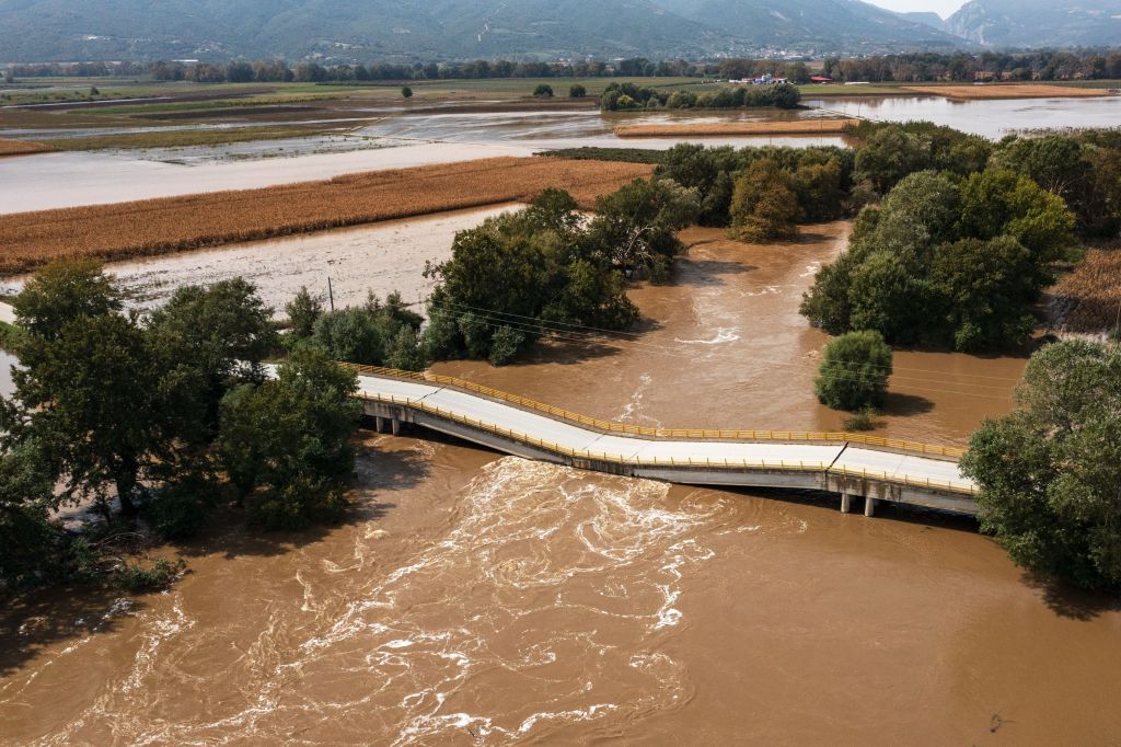 Aerial view of a collapsed bridge over a swollen, muddy river, with flooded fields and trees in the background.