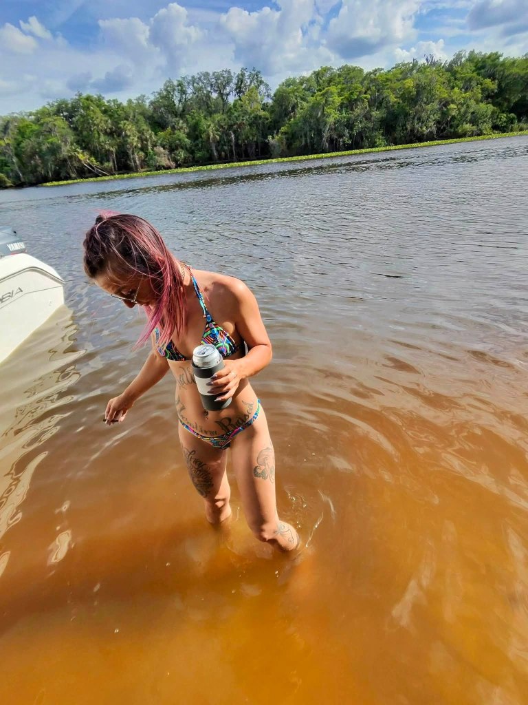 A woman with pink hair and tattoos on her arms and legs stands in brown river water, holding a drink.