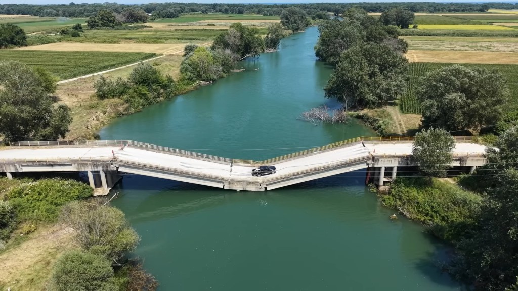 Aerial view of a black pickup truck driving over a storm-damaged bridge with a significant buckle in the middle.