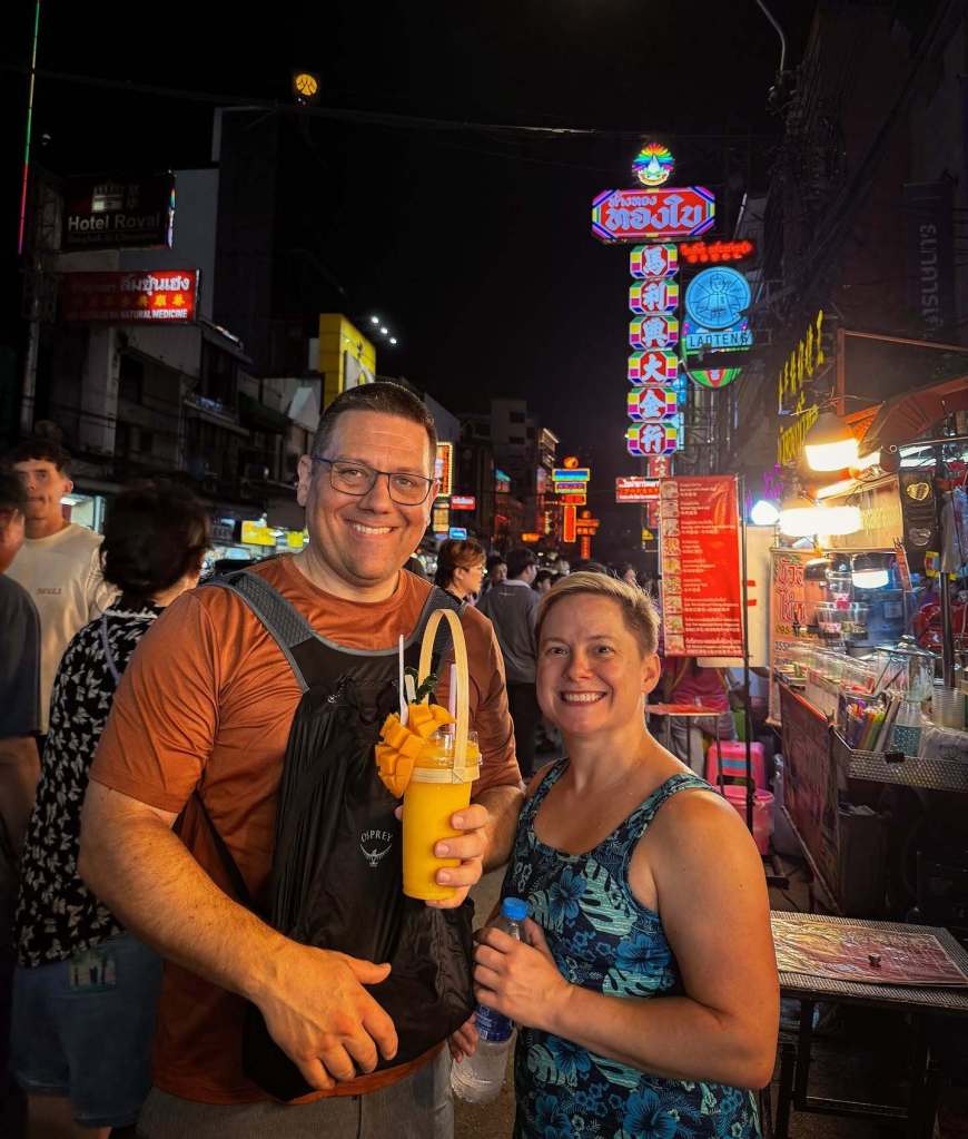 Elizabeth "Liz" Waddell and her husband, Cailen, smiling with drinks in front of street vendors and neon signs at night.