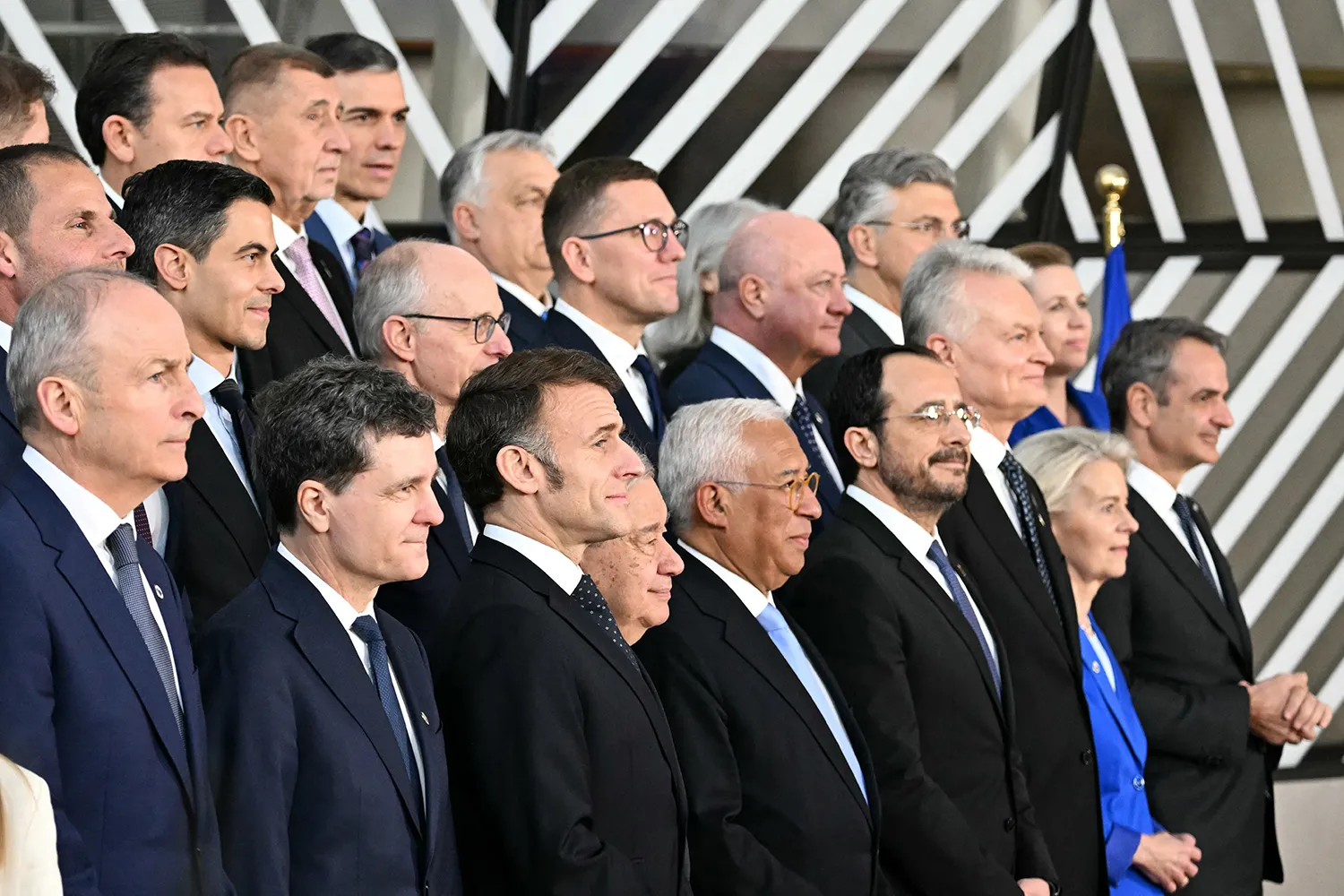 A group of people wearing dark suits and formal attire stand together on tiered steps for a group photograph. Most are looking off toward the right side of the frame in profile or three-quarter view. In the background, a wall features a bold pattern of diagonal black and white stripes.