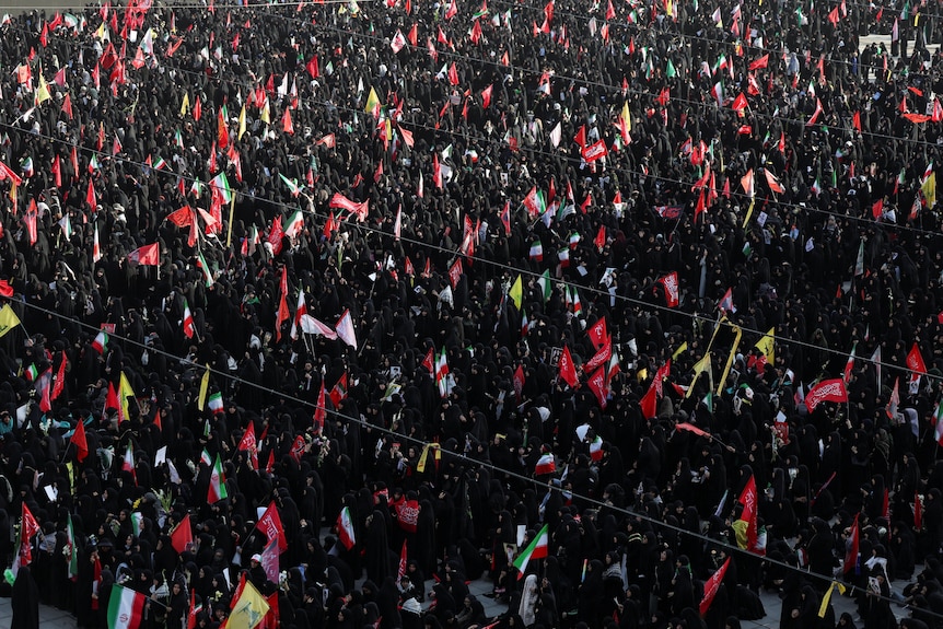 A crowd of thousands of people wearing black seen from above, the only pops of colour red and green from Iranian flags.