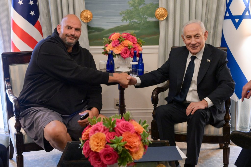 John Fetterman and Benjamin Netanyahu shake hands while seated, with flags of the U.S. and Israel in the background.