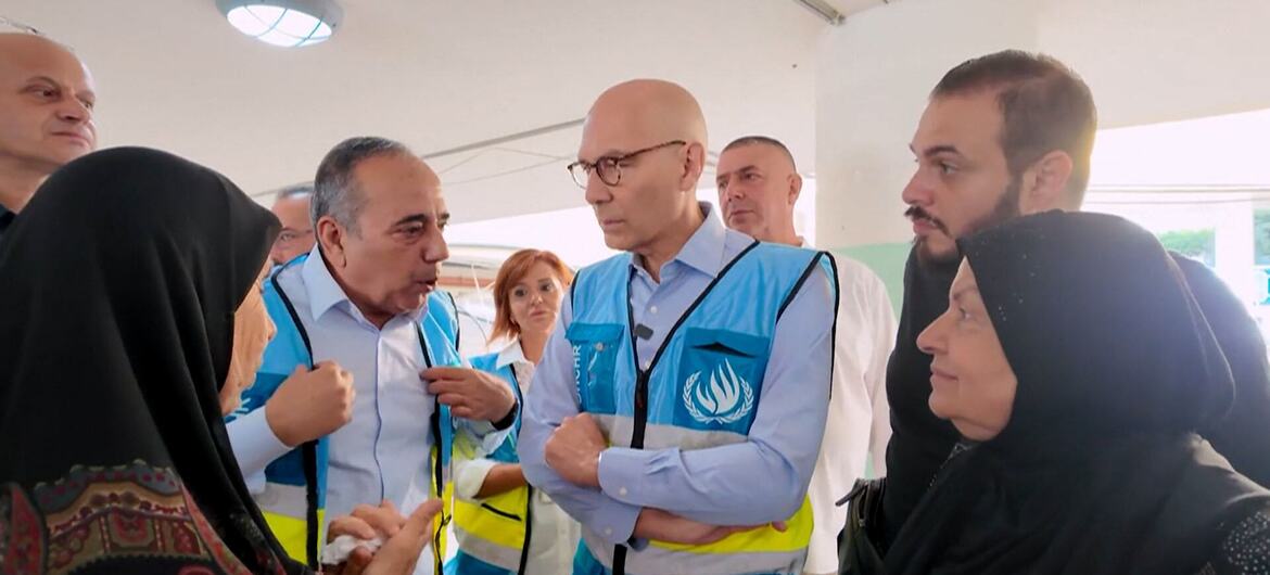 A group of Middle Eastern men and women, some wearing blue vests with the UNRWA logo, are gathered and engaged in a discussion.