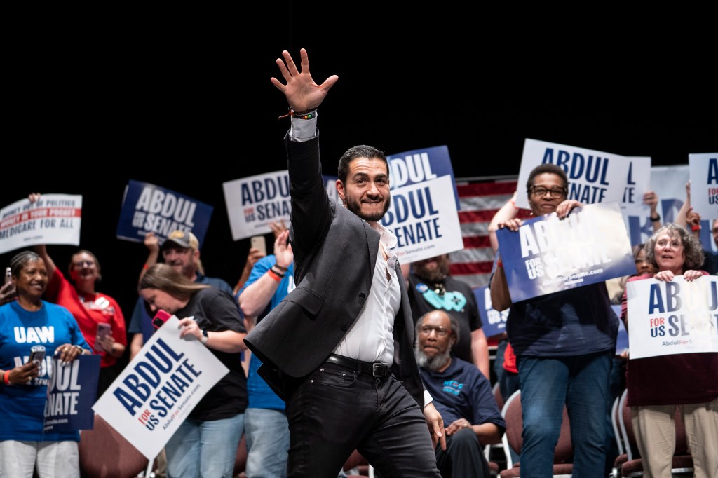Abdul El-Sayed waves to the crowd at a rally, surrounded by supporters holding "Abdul for US Senate" signs.