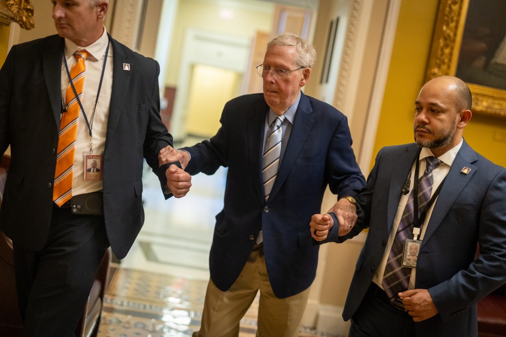 Senator Mitch McConnell, wearing glasses and a suit, is escorted through a hallway by two men in suits.