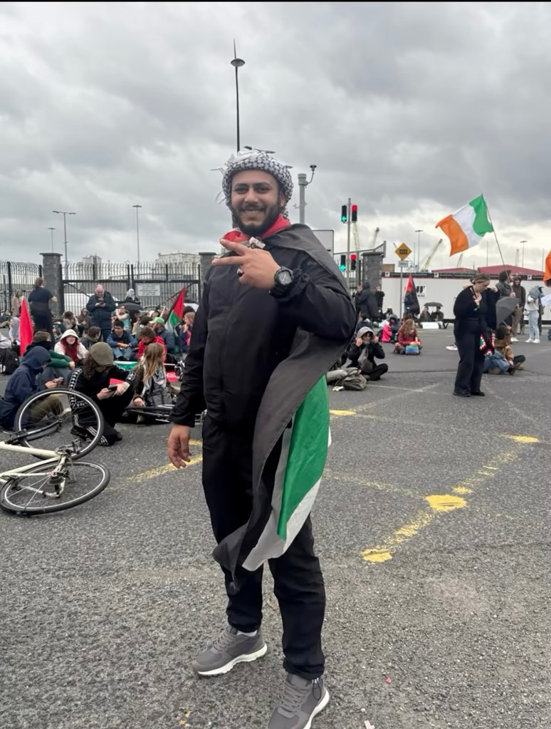 Ahmad Alsaqer at an anti-war protest, wearing a kufiya and a Palestinian flag draped over his shoulders, making a peace sign.