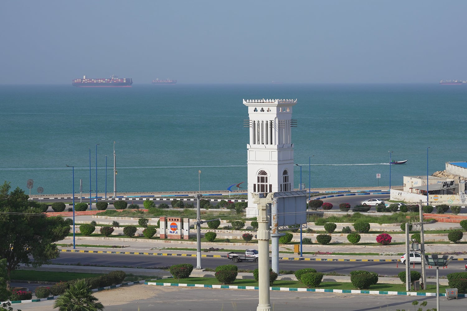 High-angle view of a coastal road and park area featuring a white tower, with large cargo ships anchored in the turquoise sea in the background.