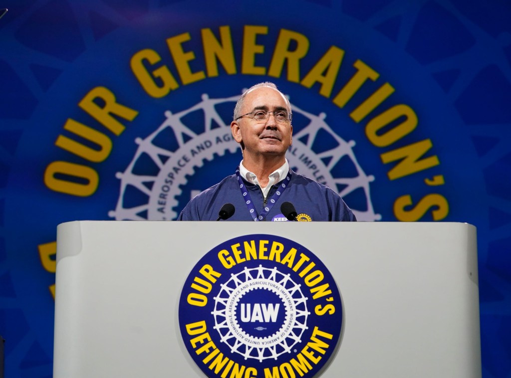 UAW President Shawn Fain speaks at a podium with the UAW logo and "OUR GENERATION'S DEFINING MOMENT" on the backdrop.