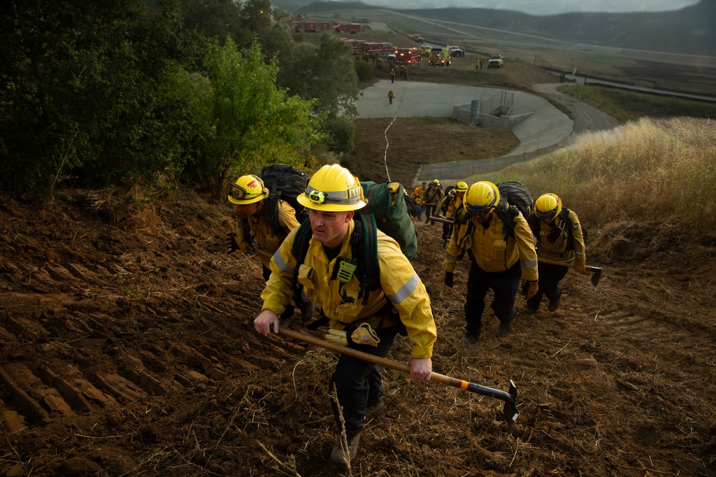 Firefighters carrying tools and gear climb a steep hillside during a wildfire response.
