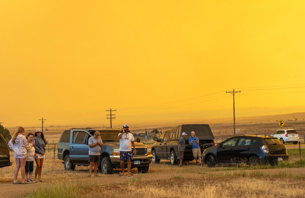 People stand beside parked vehicles under an orange, smoke-filled sky during a wildfire.