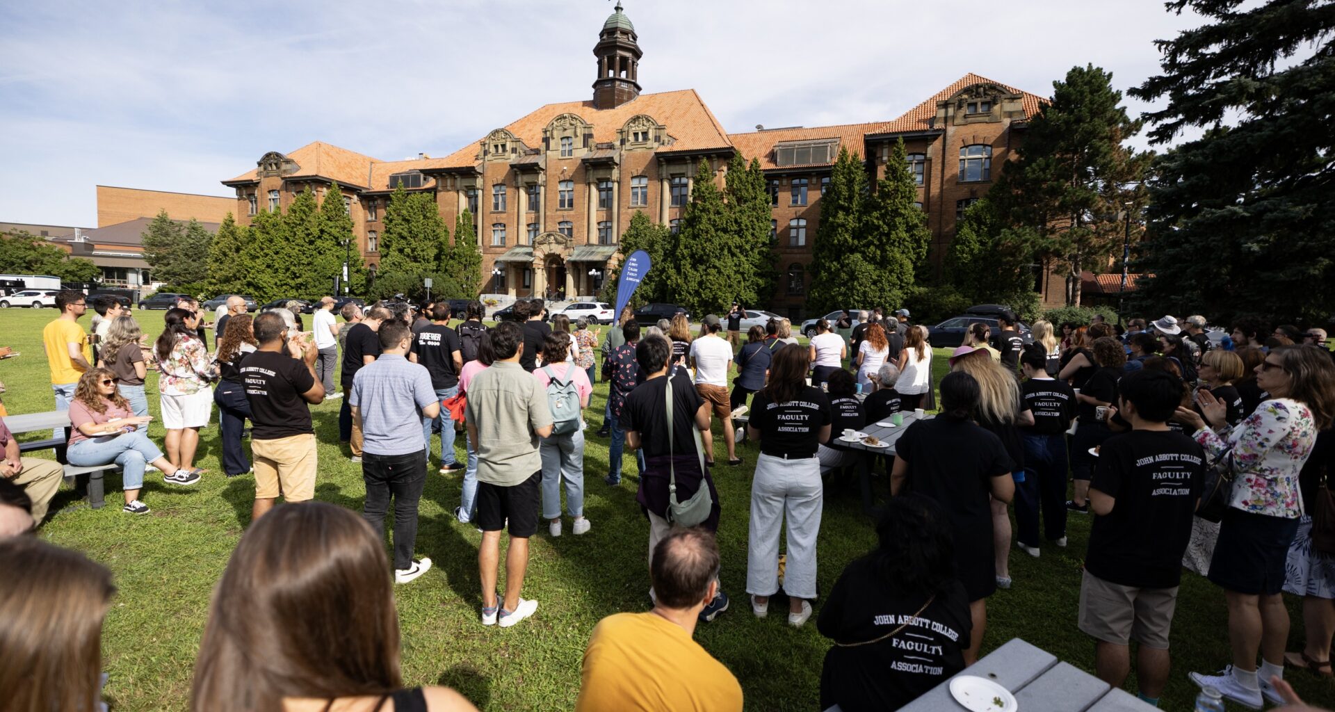 A group of people gather on the lawn of a college, shown in the background.