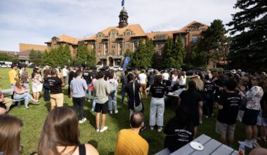 A group of people gather on the lawn of a college, shown in the background.