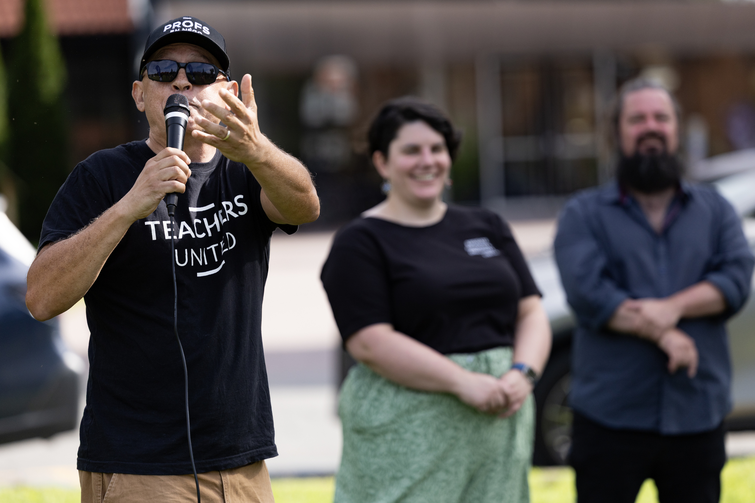Two people watch a third speak into a hand-held microphone outside.