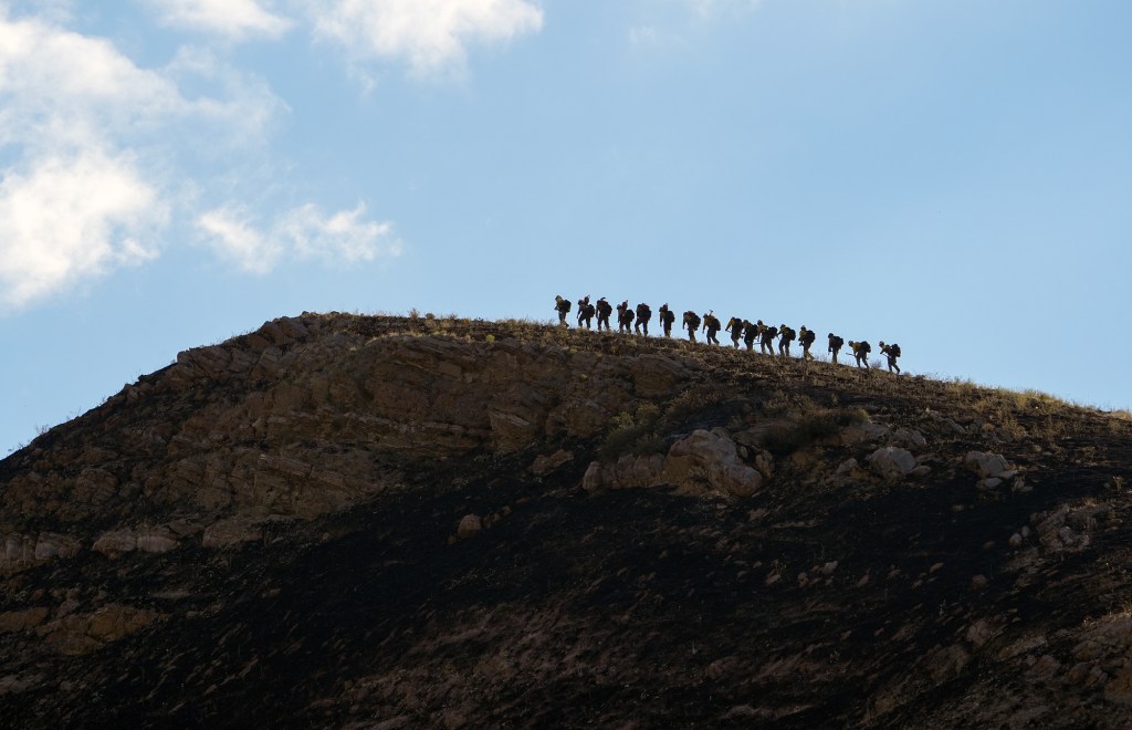 A line of firefighters hikes along the ridge of a rocky, fire-scorched hillside.