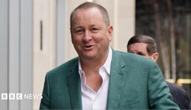 Businessman Mike Ashley wearing a white shirt, and green suit jacket, smiling at the camera as he walks with a man behind him