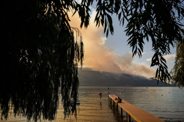A woman jumps off a dock into Okanagan Lake, in Lake Country, British Columbia, as the Quilpituk Creek wildfire burns on the mountainside across the lake near Fintry, Aug. 3, 2026. (Darryl Dyck/The Canadian Press via AP, File)