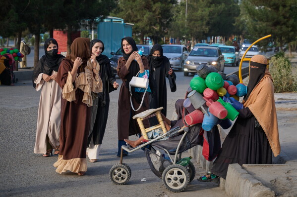 A group of young women walk past a woman selling plastic water jugs along a street on the eve of the fifth anniversary of the U.S. withdrawal and the start of Taliban rule in Kabul, Afghanistan, Friday, Aug. 14, 2026. (AP Photo/Nava Jamshidi)