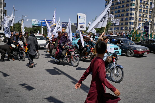 A boy walks past Taliban members riding motorcycles and waving flags during celebrations marking the fifth anniversary of the U.S. withdrawal from Afghanistan and the Taliban's return to power, in Kabul, Afghanistan, Saturday, Aug. 15, 2026. (AP Photo/Nava Jamshidi)