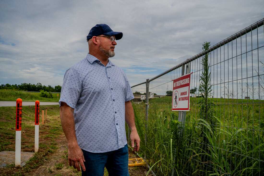 A man wearing a baseball cap stares into a lot with tall grass and a fence that says "no trespassing."