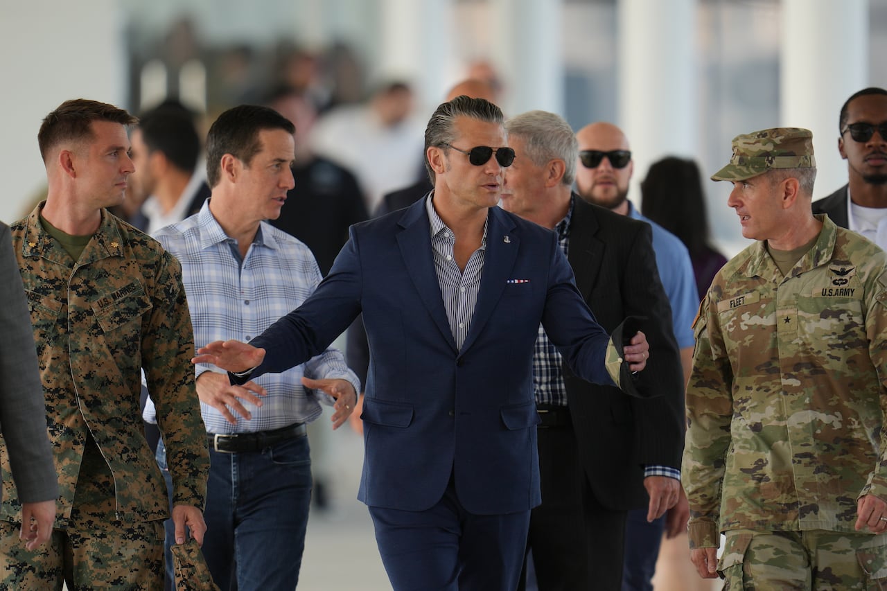 A man in sunglasses and a suit jacket and collared shirt stands in between military members in camouflage uniforms.