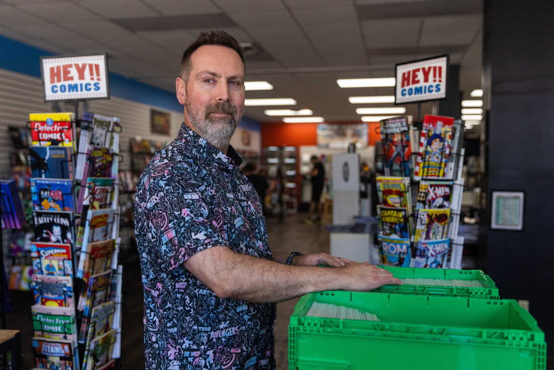 Marc Bowker, owner of Alter Ego Comics in Lima, Ohio, thumbs through a green bin of comic books and either side of him are two shelves with signs that read "Hey!! Comics." He has short brown hair and a graying beard. He is wearing a patterned button up shirt that has "Avengers" written on it along with Marvel characters such as Iron Man and Captain America.