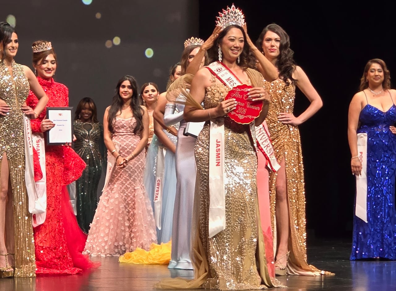 A woman wearing a sparking gown stands on a stage as a sparking crown is placed on her head. Other women in gowns stand in the background.