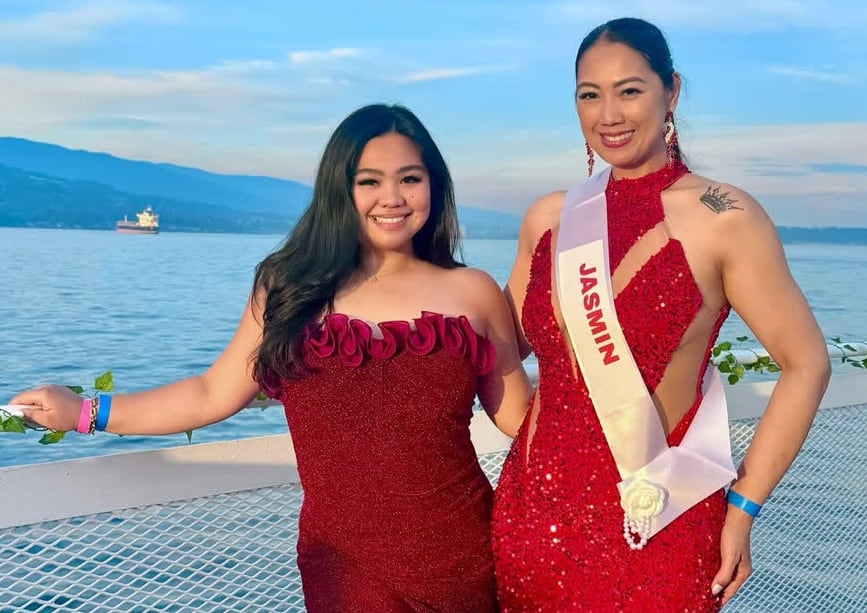 Two women in red cocktail gowns stand by a railing with water and  hills in the distance behind them. One of them is wearing a white pageant sash.