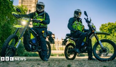 Two Metropolitan Police officers in full riding gear and helmets sit on high-powered Sur-Ron police e-bikes in a park, with the Houses of Parliament visible in the background under a clear blue sky.