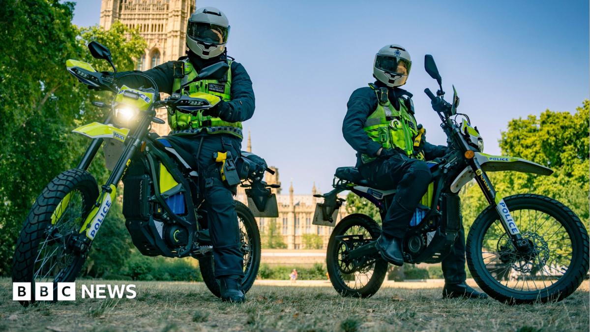 Two Metropolitan Police officers in full riding gear and helmets sit on high-powered Sur-Ron police e-bikes in a park, with the Houses of Parliament visible in the background under a clear blue sky.