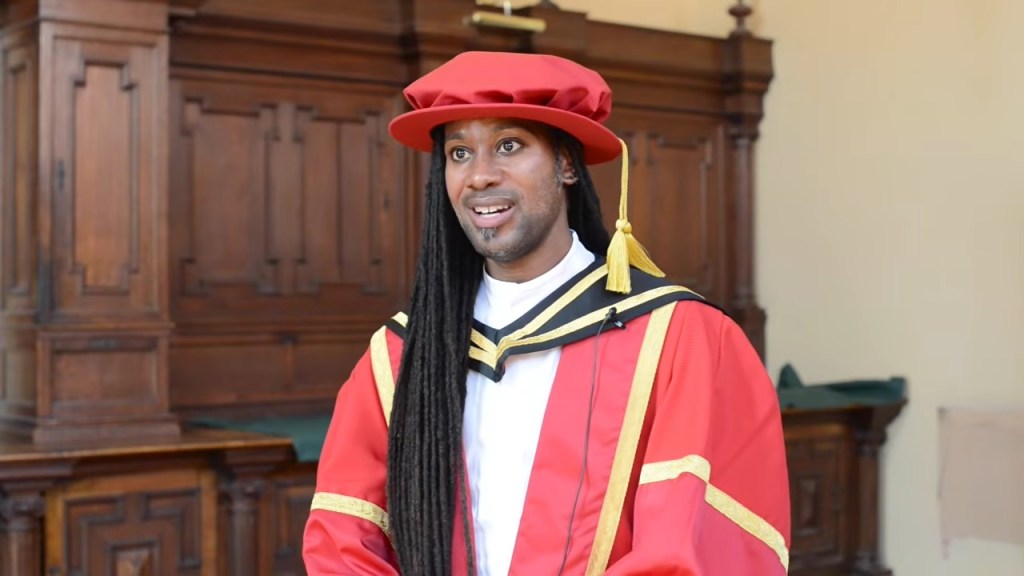 Jason Arday in academic regalia, wearing a red cap and gown, in an indoor setting.
