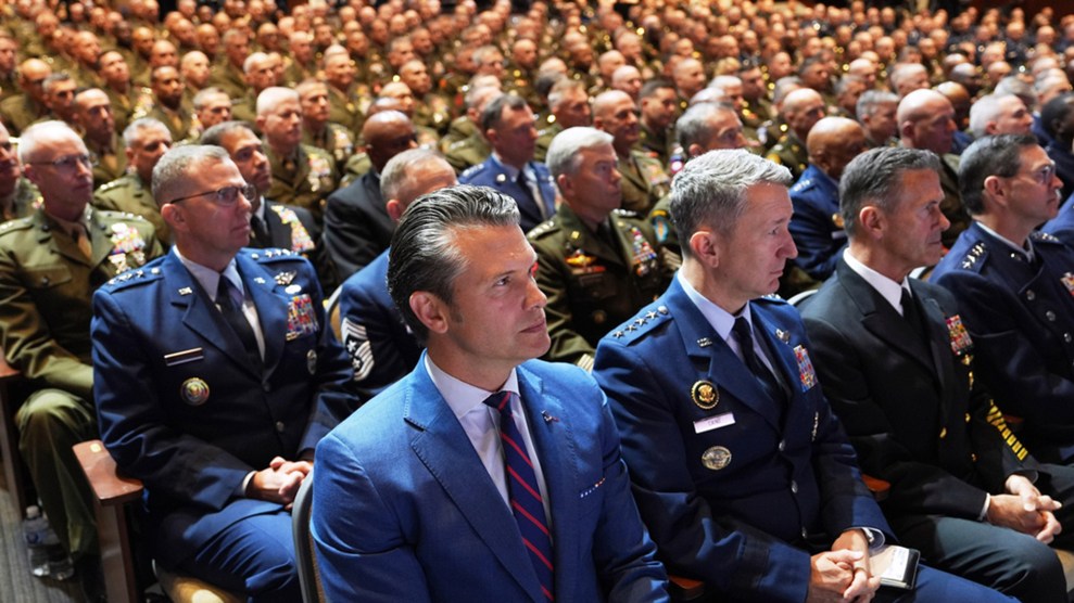 Defense Secretary Pete Hegseth, in a blue suit and striped tie, sits in an auditorium surrounded by rows of senior U.S. military officers in dress uniforms from multiple service branches.