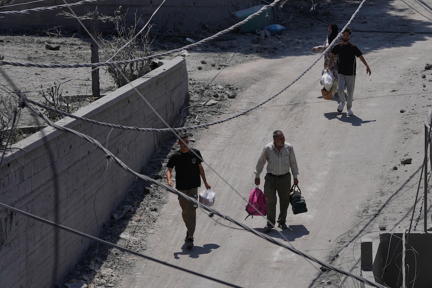 Three men walk an unsealed road carrying multiple bags as they leave an area which has been air raided