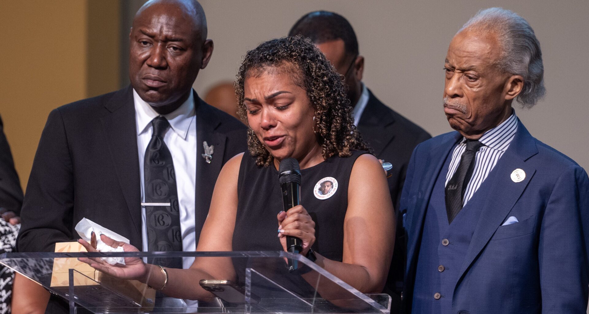 A woman speaks emotionally at a podium, holding tissues, flanked by two men in suits during a solemn event.