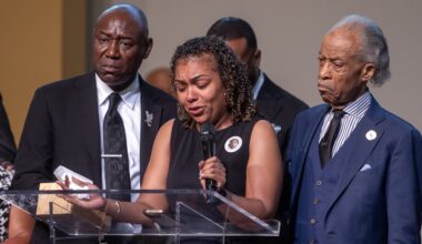 A woman speaks emotionally at a podium, holding tissues, flanked by two men in suits during a solemn event.