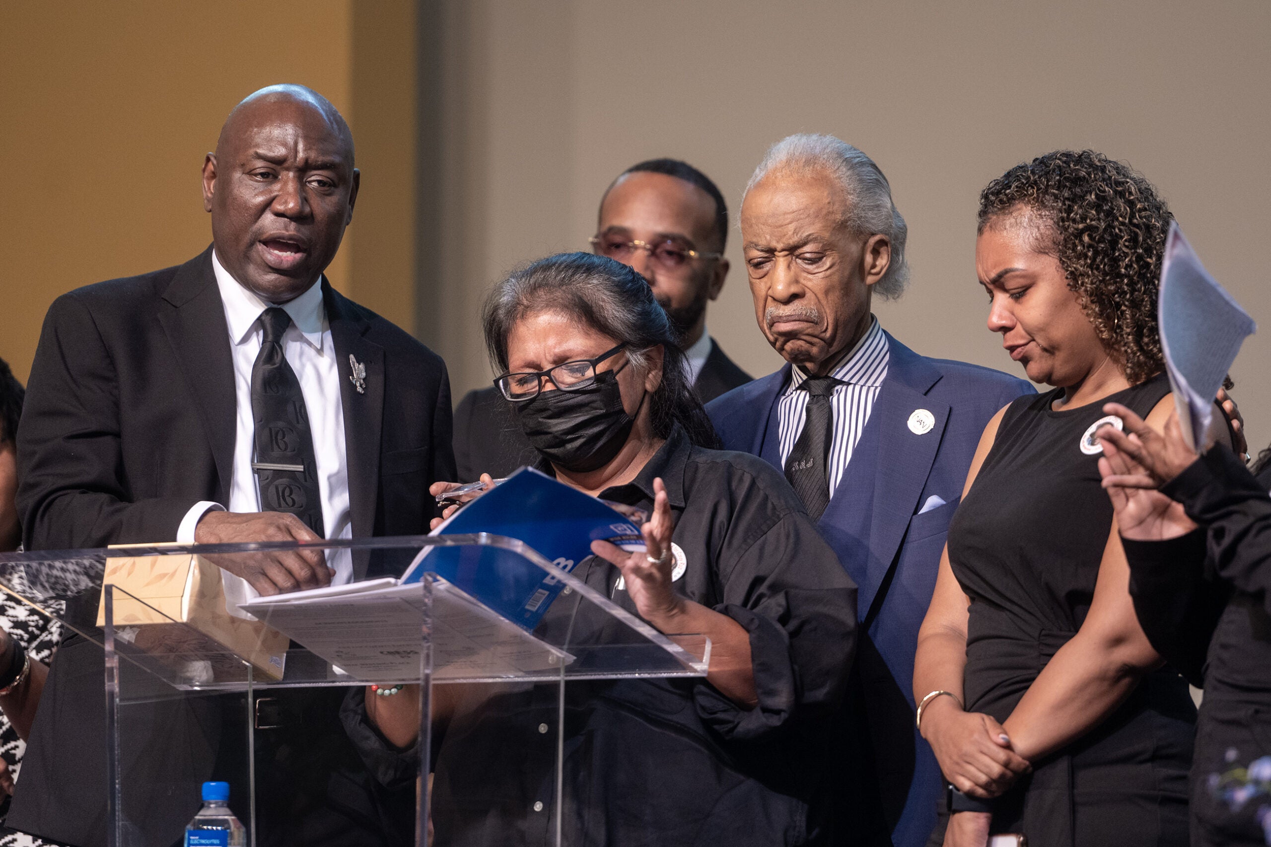 A group of people stands together at a podium, with some appearing emotional. One person reads from a document while others look on.