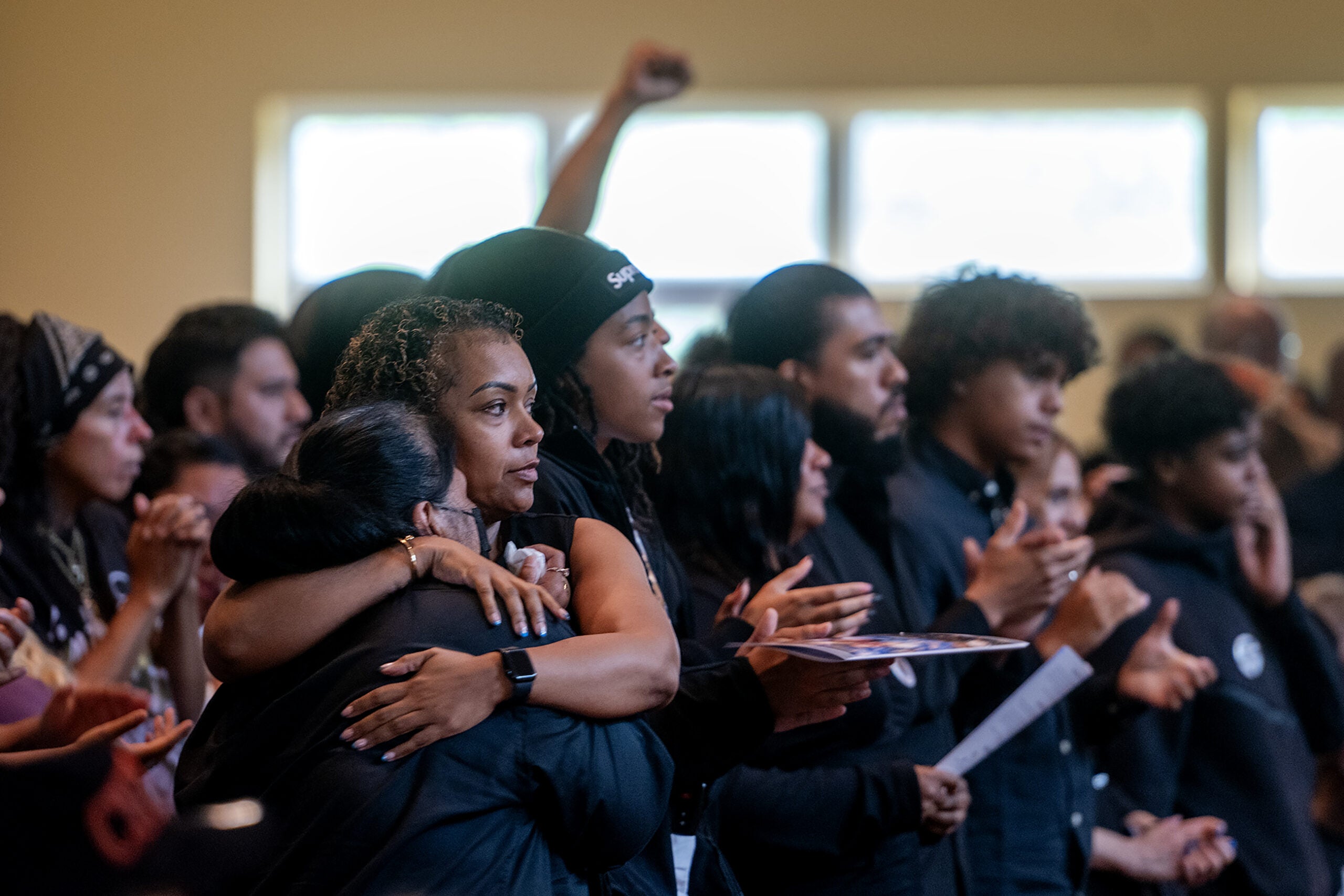A group of people dressed in dark clothing stand close together indoors, some hugging and others clapping, with serious and emotional expressions.