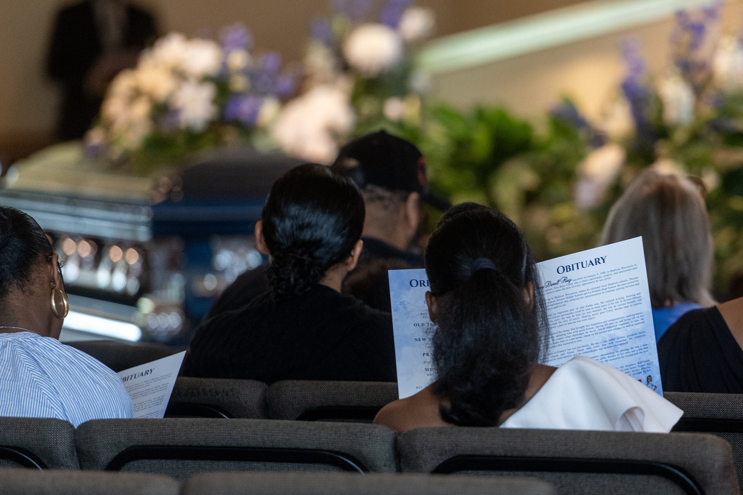 People seated at a funeral service read printed obituaries; a casket and floral arrangements are visible at the front of the room.